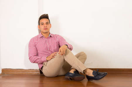 Hispanic male wearing red white shirt sitting on wooden floor against wall empty background.の写真素材
