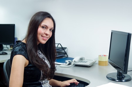 Hispanic brunette office woman sitting by desk and working on computer with positive attitude smiling.の写真素材