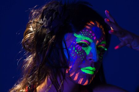 Headshot woman wearing awesome glow in dark facial paint, blue based with other neon colors and obscure abstract background, facing camera.の写真素材