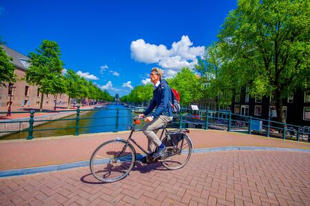 Amsterdam, Netherlands - July 10, 2015: Typical Dutch man bicycling over a channel bridge, on his way to work, beautiful blue sky.のeditorial素材