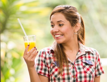 Young woman in square pattern shirt enjoying a yellow juice, garden environment.の写真素材