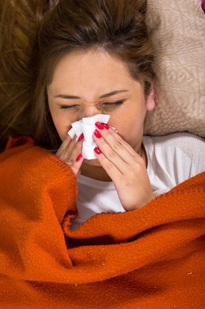Brunette lying down under orange blanket and blowing her nose, sick with flu concept.の写真素材