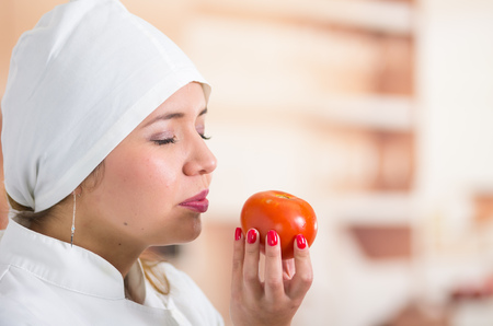 Closeup headshot of woman chef holding up a tomato and smelling it with her eyes closed.の写真素材