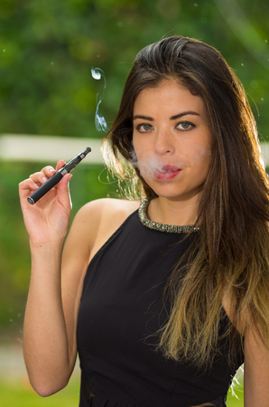 Classy brunette lady wearing black dress standing in garden environment smoking e-cigarette and posing for camera.の写真素材