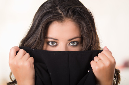 Headshot attractive brunette facing camera covering half her face with black clothing, white studio background.の写真素材