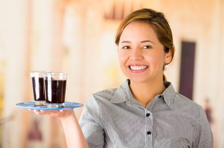 Young brunette waitress wearing uniform with friendly smile, holding up tray containing two glasses of dark liquid using one arm.の写真素材