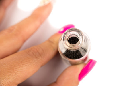 Closeup fingers holding small glass bottle with dark colored nail polish inside, white surface...