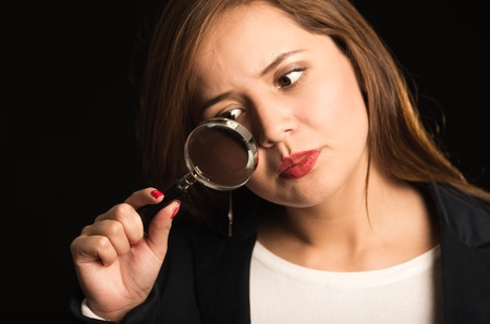 Young woman holding magnifying glass in front of face, slightly tilting head forward looking through it, black background.の写真素材