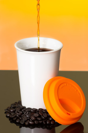 Great image showing stream of coffee pouring into white cup from above with coffe beans on surface sourrounding mug and orange lid sunset colored background.の写真素材