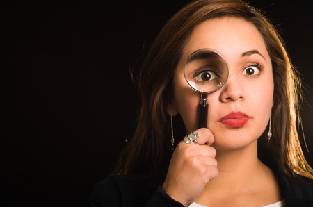 Young woman facing camera holding magnifying glass over right eye creating enhanced effect, black background.の写真素材