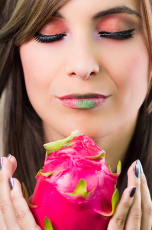 Headshot brunette, dark mystique look and green lipstick, holding up pink pitaya fruit with both hands facing camera.の写真素材