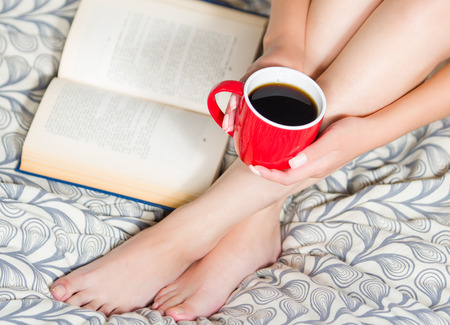 Woman sitting on bed, only legs visible, holding red coffee cup and open book lying next to feet.の写真素材