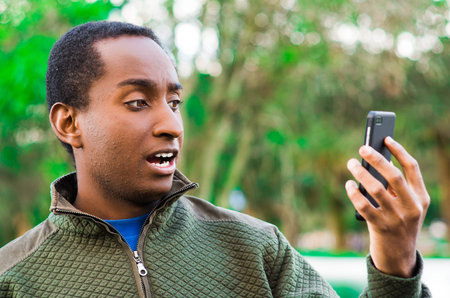 Handsome hispanic black man wearing green sweater in outdoors park area holding up phone and...