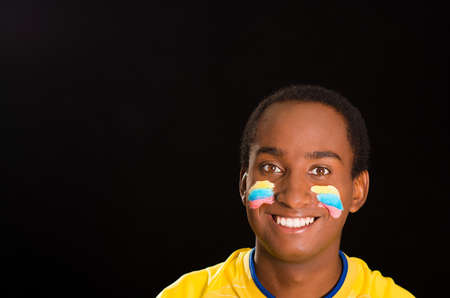 Headshot dark skinned male wearing yellow football shirt in front of black background, flag facial paint, facing camera and smiling happily.の写真素材