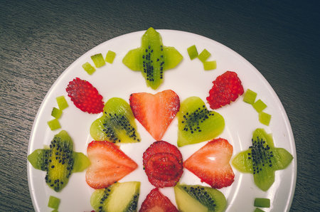 Sliced kiwi and strawberries lying in neatly placed pattern on white plate, as senn from above.