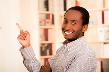 Man wearing casual clothes posing happily for camera, index finger pointing upwards, bookshelves background.の写真素材