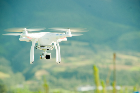 White drone with camera mounted underneath hovering midair, mountain landscape background.の写真素材