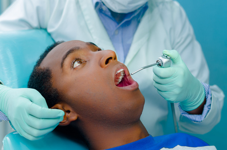 Young hispanic male lying in dental chair looking up, dentists hands with gloves holding tool into patients mouth.の写真素材