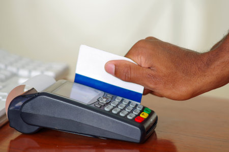 Closeup hand swiping credit card through payment terminal device sitting on wooden surface.