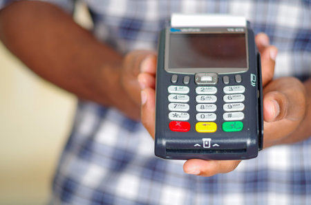 Man wearing blue white square pattern shirt holding up credit card payment terminal in front of...