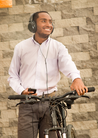 Man wearing grey office pants, white red business shirt standing by bicycle holding mobile phone, headphones on head, smiling and posing, brick wall background.の写真素材