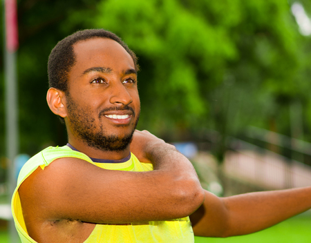 Man wearing yellow shirt stretching arms in park sorrounded by green grass and trees, training concept.の写真素材