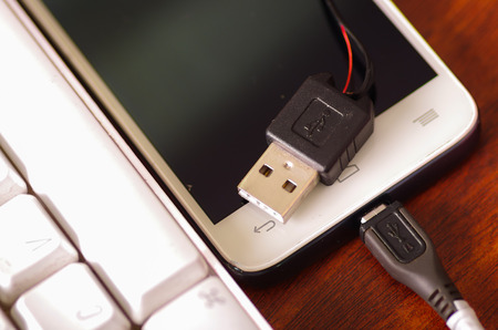 Broken usb mobile charger cable connected into smartphone lying on wooden surface next to computer keyboard, part of wire exposed through crack in plastic protection.の写真素材