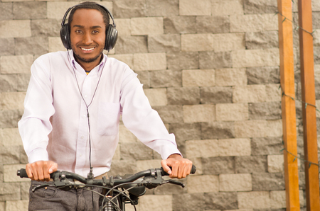 Man wearing grey office pants, white red business shirt standing by bicycle holding mobile phone, headphones on head, smiling and posing, brick wall background.の写真素材