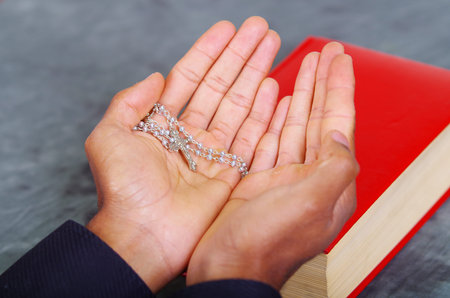 Red bible lying on blue desk with mans open hands above book, rosary cross placed inside, religion...