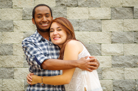 Interracial charming couple wearing casual clothes posing for camera and embracing in front of grey brick wall.の写真素材