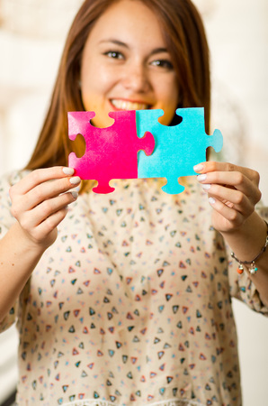 Headshot charming brunette woman holding up big puzzle pieces in pink and blue, smiling happily to camera, white studio background.の写真素材