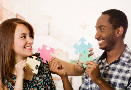 Interracial charming couple holding up large puzzle pieces and happily interacting having fun, blurry studio background.の写真素材