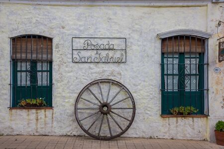 COLONIA DEL SACRAMENTO, URUGUAY - MAY 04, 2016: sign hanging from a white old wall with two nice ancient green windows and a cart wheel under the sign.のeditorial素材