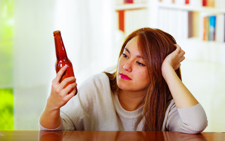 Woman wearing white sweater sitting by bar counter lying over desk staring at beer bottle, drunk depressed facial expression, alcoholic concept.の写真素材