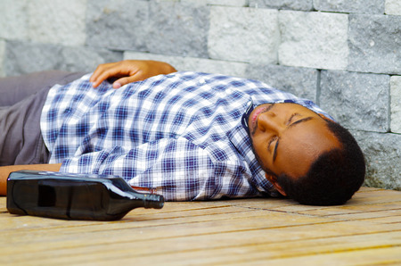 Man wearing casual clothes lying drunk passed out on wooden surface next to grey brick wall, empty bottle beside him.の写真素材
