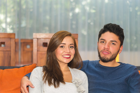 Young charming couple seated and embracing in orange sofa smiling to camera, hostel environment.の写真素材