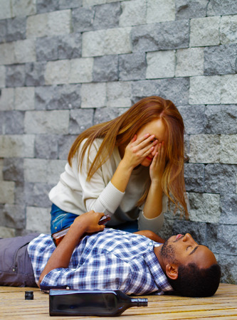 Man wearing casual clothes lying drunk passed out on wooden surface, pretty woman sitting beside him trying to get contact by touching and shaking.の写真素材