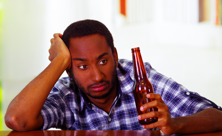Handsome man wearing white blue shirt sitting by bar counter lying over desk holding brown beer bottle, drunk depressed facial expression, alcoholic concept.の写真素材
