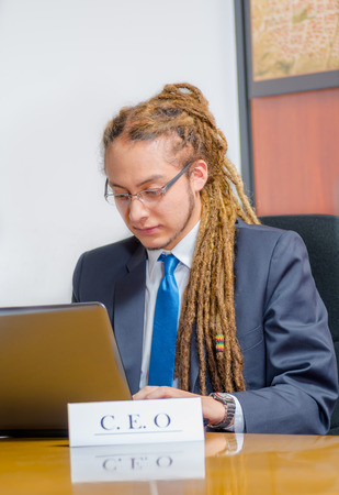 Handsome man with dreads and business suit sitting by desk looking at laptop screen working, young manager concept.の写真素材