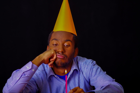 Bored looking man wearing blue shirt and hat sitting by table holding a single candle burning, sad expression facing camera, celebrating alone concept.の写真素材