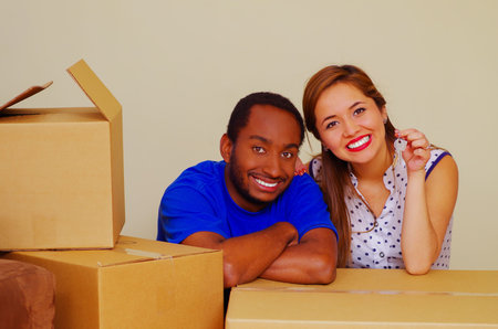 Charming interracial couple smiling to camera while leaning on cardboard boxes, moving in concept.