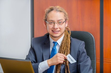 Handsome man with dreads, glasses and business suit sitting by desk placing a paper which has the word ceo written on it in his pocket, young executive concept.の写真素材
