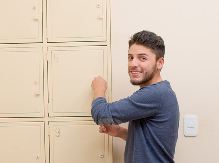 Young handsome man wearing blue sweater smiling and opening metal locker door, rack of lockers stacked.の写真素材