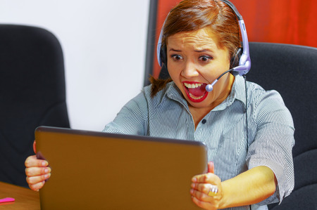 Young attractive woman wearing office clothes and headset sitting by desk looking at computer screen, grabbing laptop, upset body language screaming out loud.の写真素材