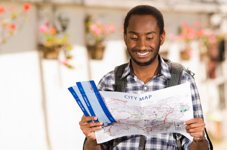Young happy man wearing casual clothes and backpack, holding travel tickets while looking at map,...