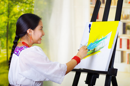 Young brunette woman wearing traditional andean clothing painting on canvas inside studio, garden window background.の写真素材