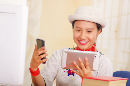 Young pretty girl wearing white shirt with colorful flower decorations and fashionable hat smiling, sitting by desk looking at tablet screen, stack of books, bright background.の写真素材