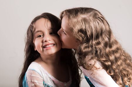 Adorable big sister kissing sibling on cheek, both wearing matcing blue dresses posing together happily, studio background.の写真素材