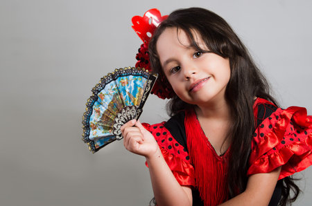 Cute little girl wearing beautiful red and black dress with matching head band, posing for camera using chinese hand fan, studio background.の写真素材