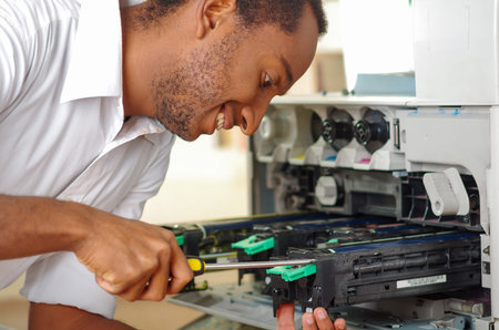 Man leaning over open photocopier during maintenance repairs using handheld tool, black mechanical...